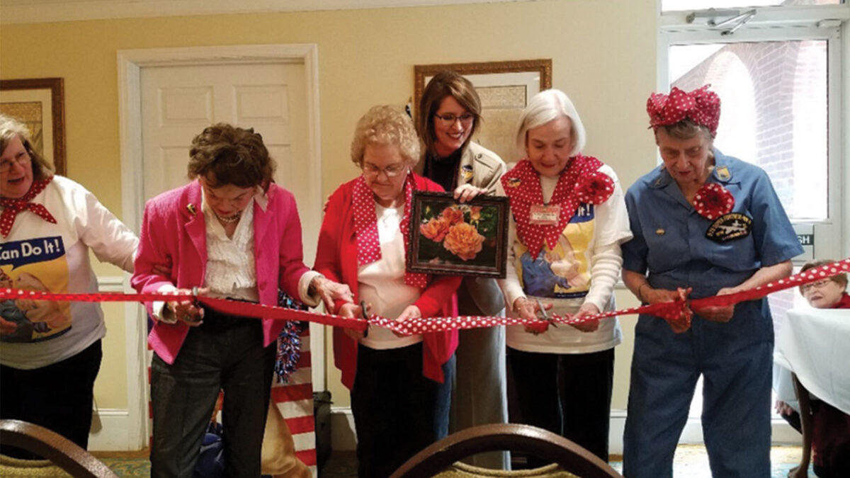 A group of six elderly women are cutting a red ribbon at an indoor event. One woman in the center holds a framed picture. They are all dressed in festive attire.