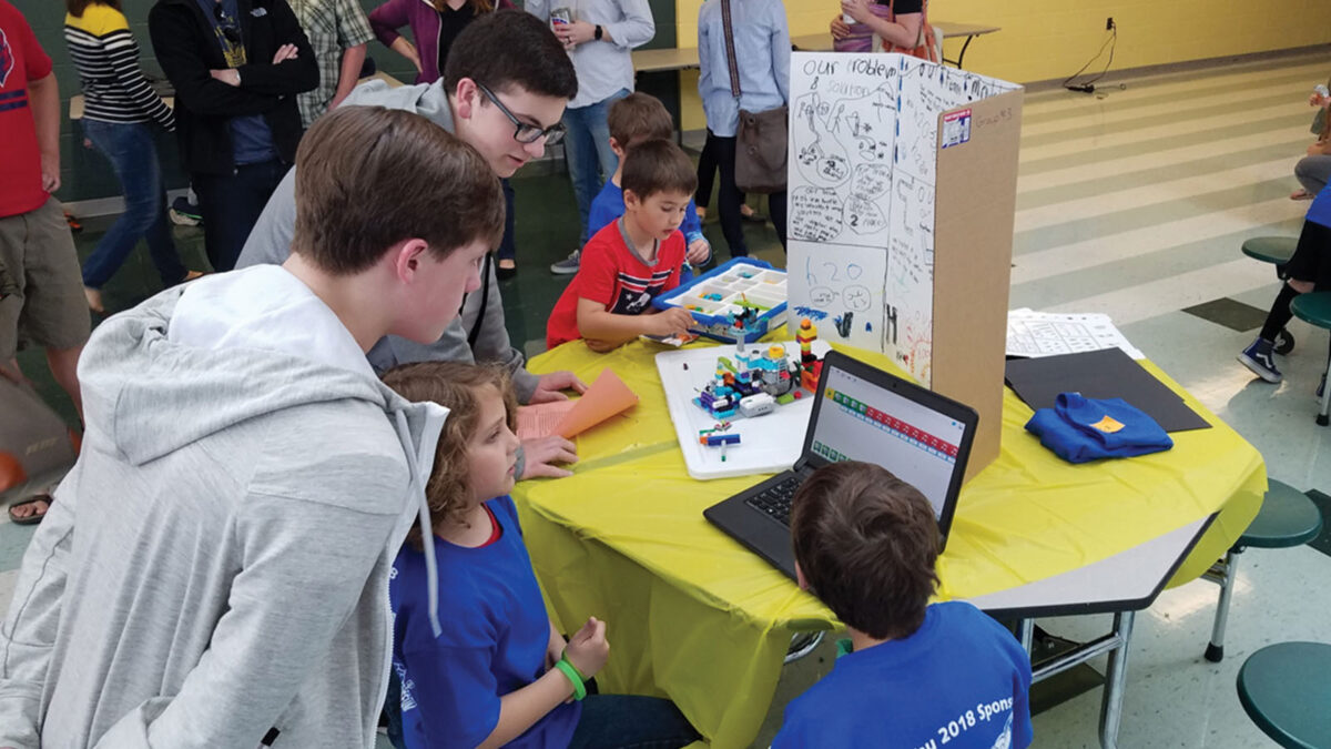 Several children and an adult gather around a table to interact with a laptop and display Lego structures at a science fair. A trifold board with notes is set up on the table.