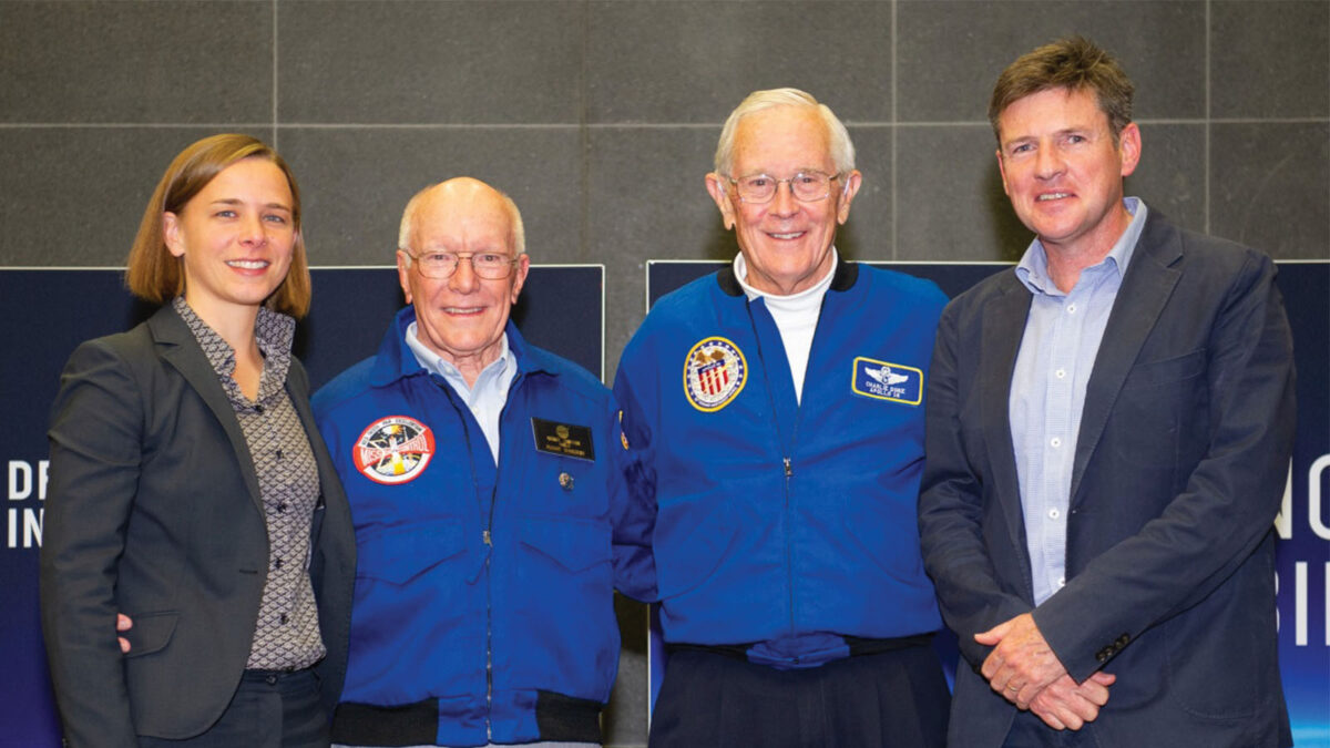 Four people are standing side by side, smiling. Two men in the middle are wearing blue jackets with patches, while the individuals on either side are dressed in formal business attire.