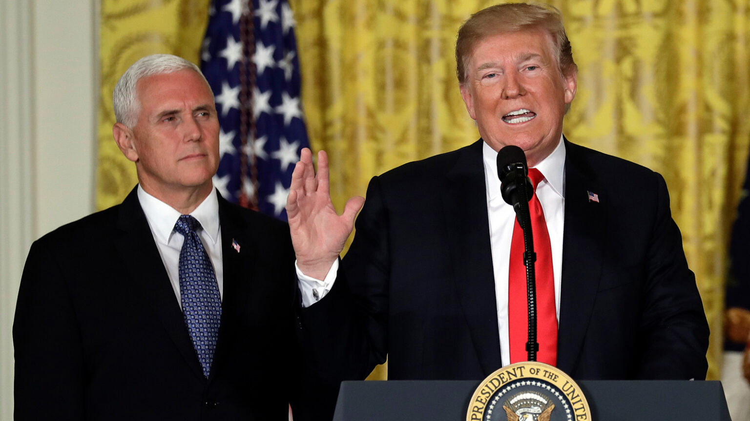 Two men in suits standing at a podium with a microphone. One man is speaking, while the other man stands with his hands clasped. An American flag is visible in the background.