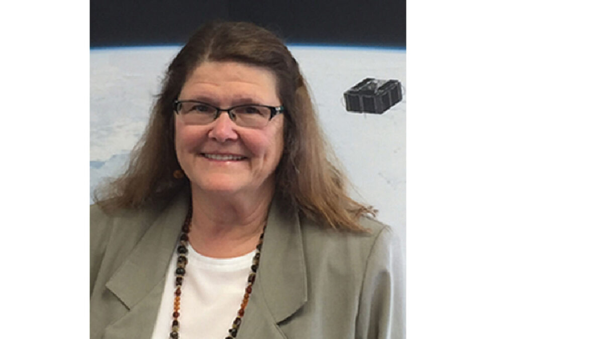 A woman with glasses and long hair wears a gray jacket and a beaded necklace, standing in front of a space-themed background.