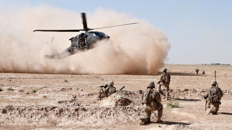 A helicopter lands in a desert area, creating a dust cloud, while military personnel in combat gear stand and crouch nearby.
