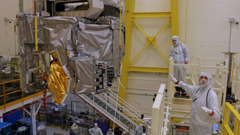 Three engineers in white cleanroom suits work on a large satellite component in a high-tech laboratory. The satellite is encased in reflective material and is supported by scaffolding and equipment.
