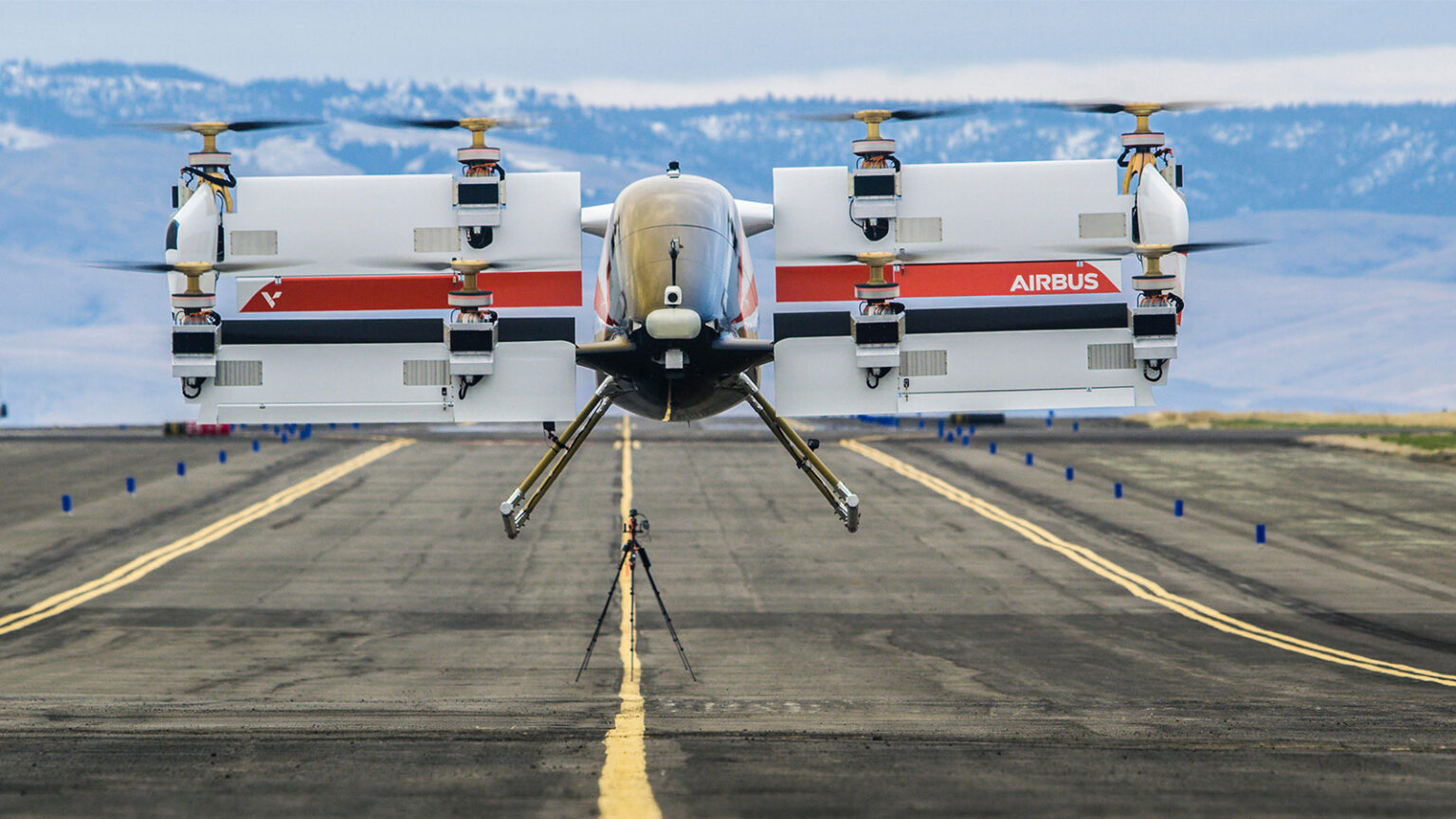 A prototype vertical takeoff and landing (VTOL) aircraft with multiple rotors hovers over a runway with mountains in the background.