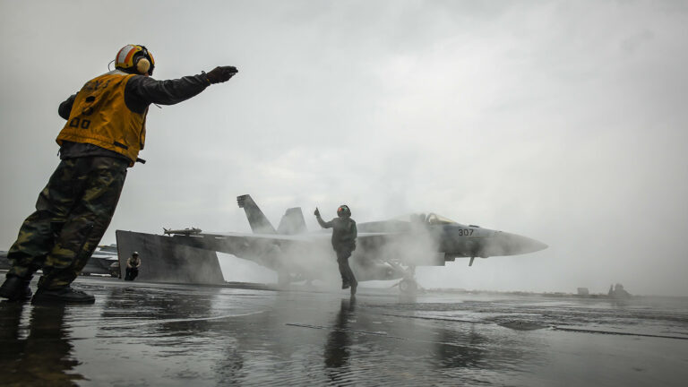 A person in a yellow uniform signals to a jet as it prepares for takeoff on an aircraft carrier. Another individual in safety gear is seen directing in front of the jet. The scene is misty and overcast.