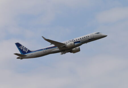 A commercial ANA (All Nippon Airways) jet aircraft is in midair, flying against a cloudy sky.