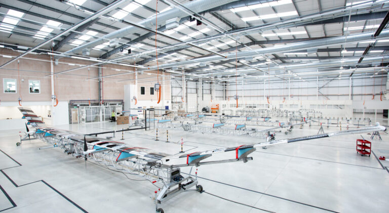 A large, open aircraft hangar with a complex, unmanned aircraft in the center. The hangar has high ceilings with numerous lights and visible structural beams.