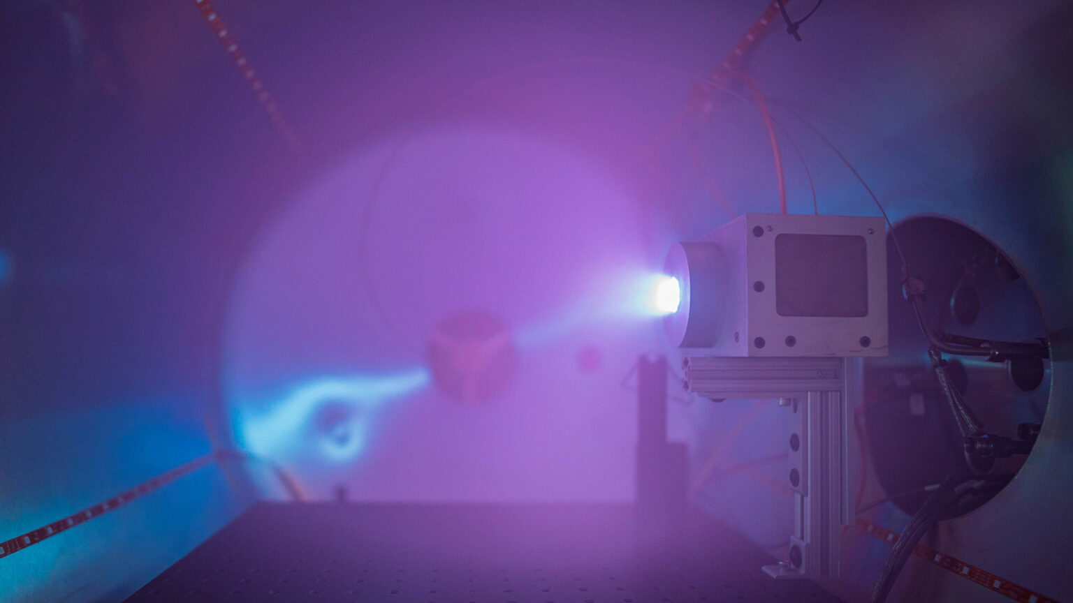 A scientific apparatus emitting a blue-purple light inside a fog-filled chamber, with various cables and components visible.