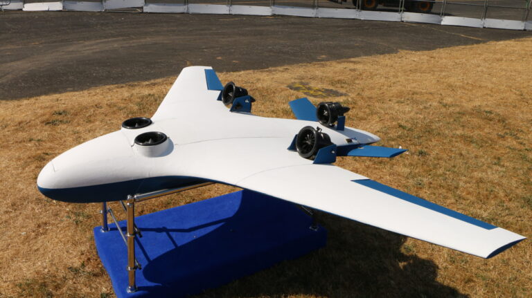 A white and blue unmanned aerial vehicle (UAV) with four propellers is displayed on a blue stand on a grassy field.