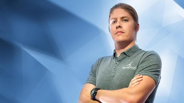 A person stands with arms crossed wearing a gray polo shirt against a geometric blue background.