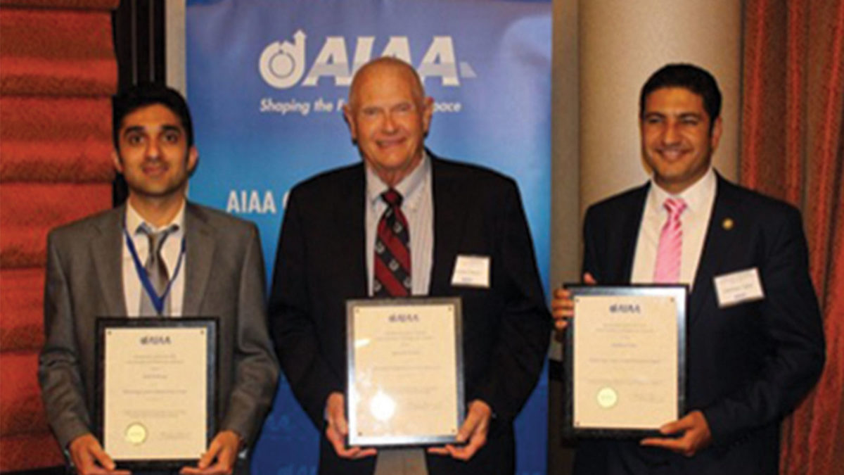 Three men holding certificates stand in front of an AIAA banner, posing for a photo and smiling.