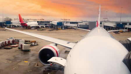An airplane is parked at an airport gate with its engine exposed. Another plane is seen in the distance with the sunset sky in the background. Luggage carts and ground equipment are visible on the tarmac.