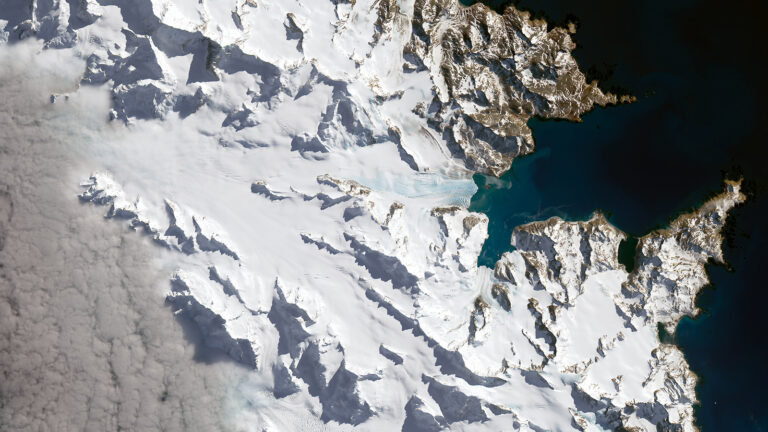 An aerial view of a snowy, mountainous landscape with rugged terrain and a dark body of water on the right side.