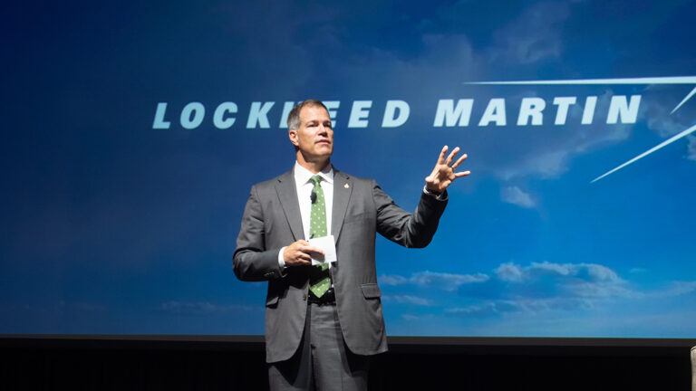 A man in a suit and tie speaks on stage in front of a screen displaying the Lockheed Martin logo.