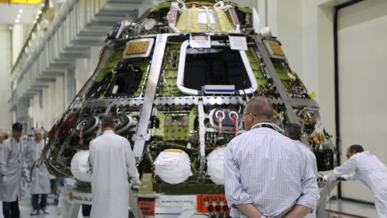 Engineers in lab coats and safety gear work on a spacecraft assembly with a person in a checked shirt observing in the foreground. The spacecraft is surrounded by various equipment and tools.