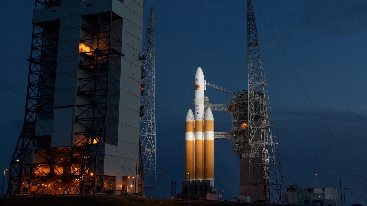 A Delta IV Heavy rocket stands on the launch pad, illuminated against the evening sky, flanked by supporting structures.