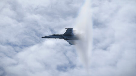 A fighter jet breaks the sound barrier, creating a visible vapor cone in the cloudy sky.