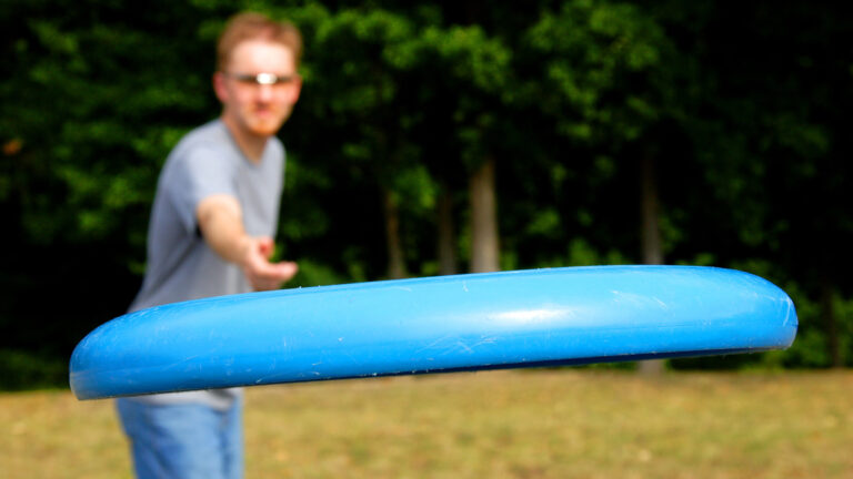 Person wearing glasses and a gray shirt is throwing a blue frisbee in an outdoor setting with trees in the background.