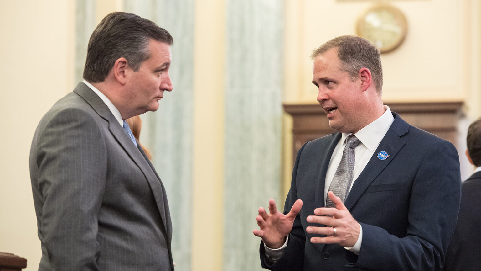 Two men in suits having a serious conversation indoors. One man gestures with his hands while speaking. A large clock is visible on the wall in the background.