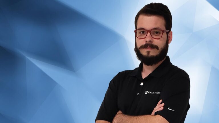 A man with glasses and a beard, wearing a black polo shirt with a logo, stands with arms crossed against a geometric blue background.
