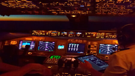 View of an illuminated airplane cockpit at night with multiple control panels and screens, pilots in uniform are seated and one holds a tablet.
