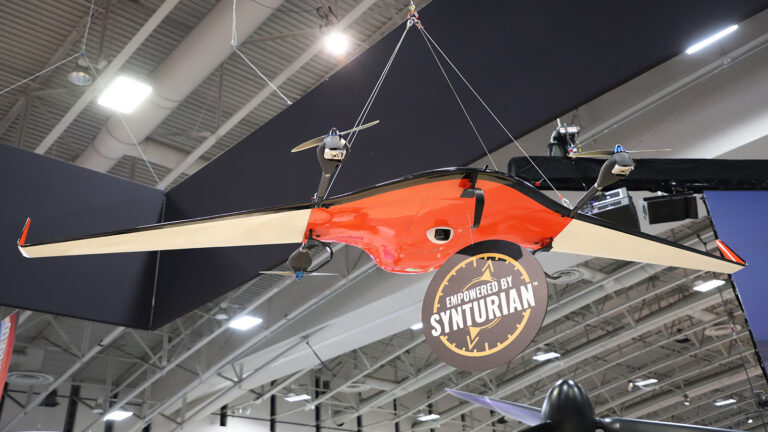 A red and black drone with a "Powered by Synturian" label is suspended from the ceiling in an industrial setting.