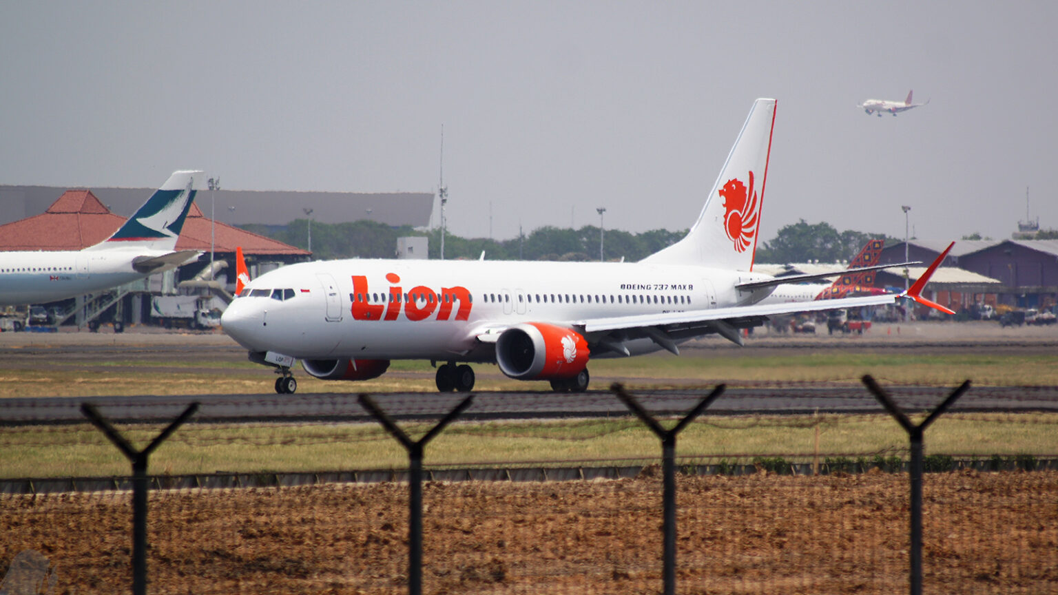 A Lion Air Boeing 737 Max 8 aircraft is taxiing on a runway at an airport, with another plane in the background and a fence in the foreground.