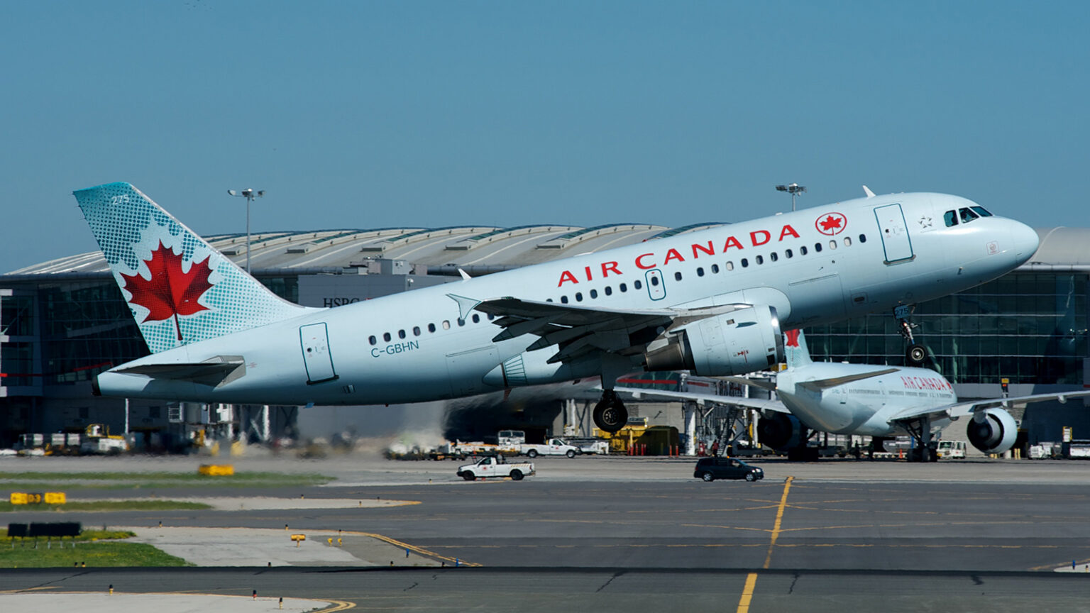 An Air Canada plane takes off from an airport runway, with another plane taxiing on the runway below, and an airport terminal in the background.