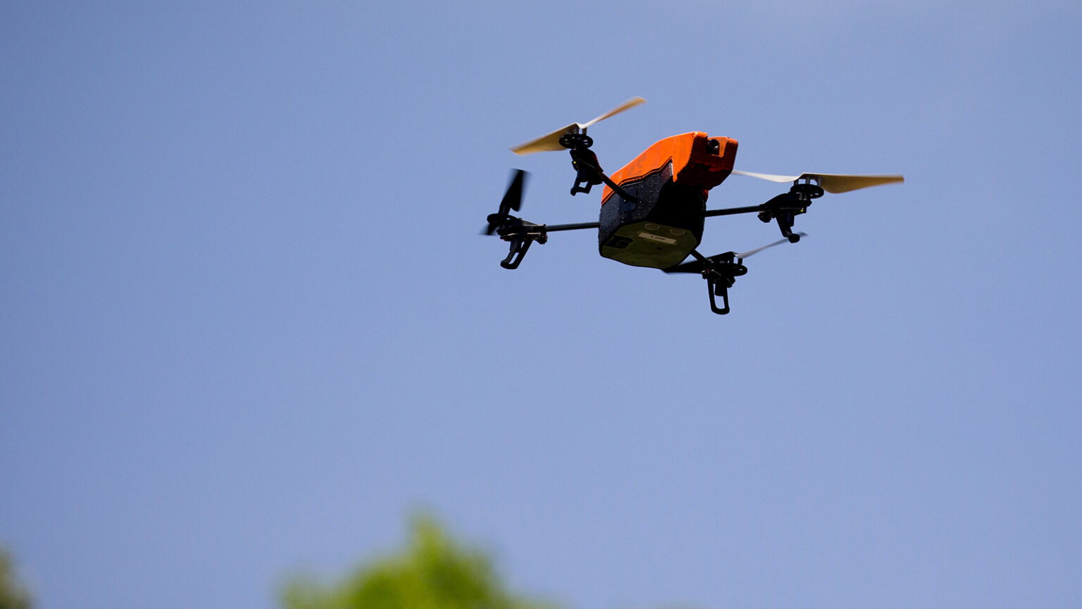 A small, four-propeller drone is flying in the sky against a clear blue background with some greenery at the bottom.
