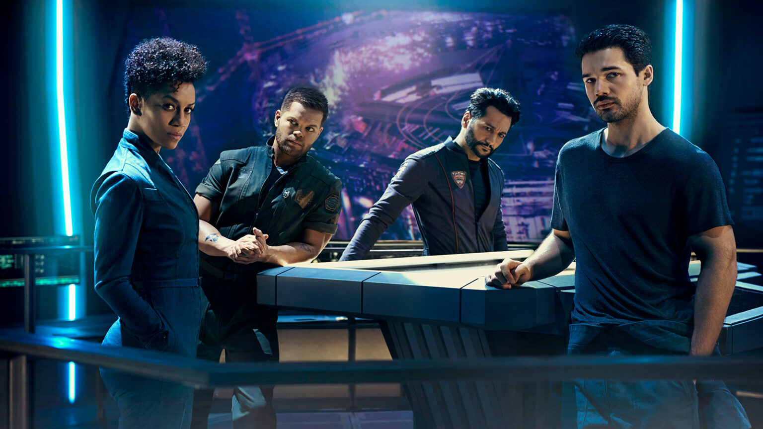 Four people stand around a futuristic-looking table in a dimly lit room with blue lighting. They are dressed in various uniforms and casual attire, appearing engaged in a serious discussion.