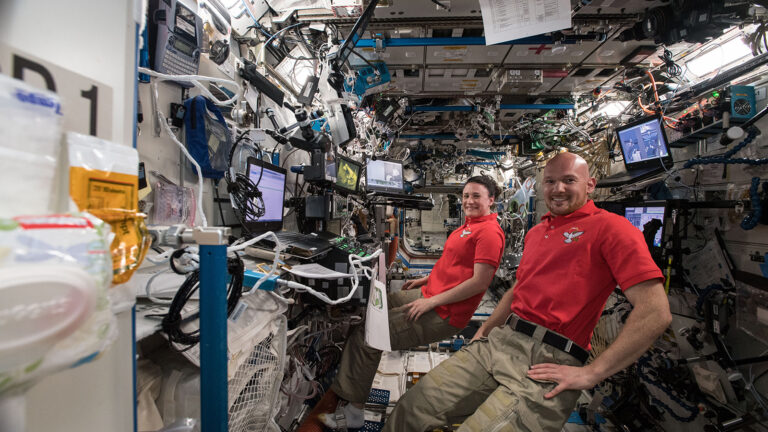 Two astronauts in red shirts work in a cluttered, equipment-filled module on the International Space Station, surrounded by screens and various instruments.