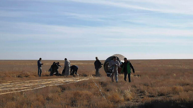 Several people stand and work around a capsule that has landed in a grassy, barren field under a blue sky.