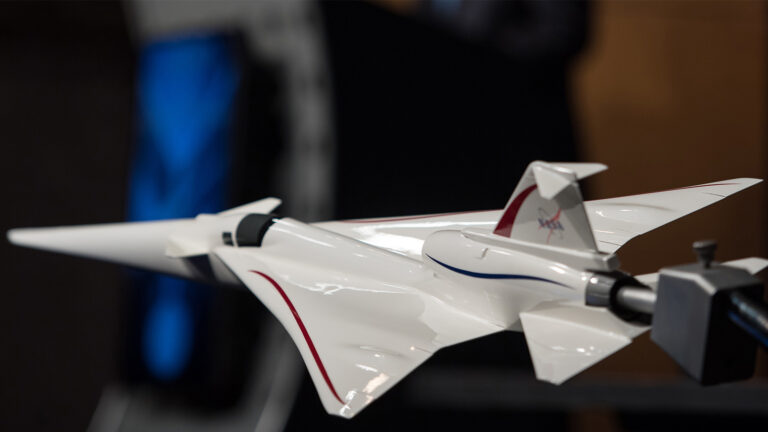 A scale model of a sleek, white supersonic aircraft displayed indoors, featuring red and blue accents and mounted on a stand for presentation.