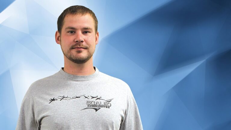 A man with short hair and a beard stands in front of a blue geometric background, wearing a gray "Scaled Composites" shirt with a plane graphic.
