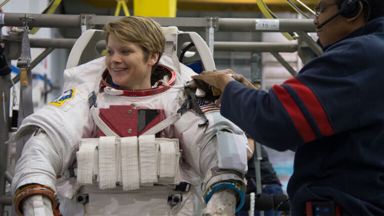 An astronaut, smiling, is being helped into a spacesuit by an assistant in preparation for a mission. They are indoors, surrounded by technical equipment.