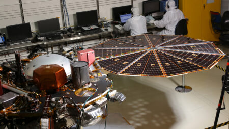 Two technicians in cleanroom suits work on computers next to a spacecraft with an extended circular solar array inside a laboratory.