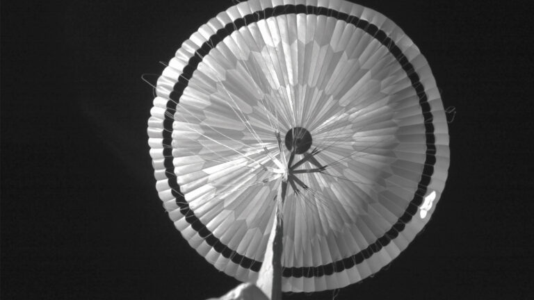 An aerial view of a deployed parachute against a dark sky. The parachute is open and radiating outward, with visible fabric lines and stitching.