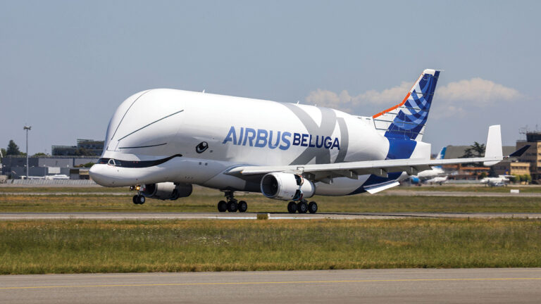 An Airbus Beluga aircraft on the runway, preparing to take off, with airport buildings and a clear sky in the background.