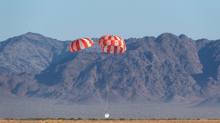 A capsule with two red and white parachutes descends in a desert landscape with a rocky mountain range in the background.