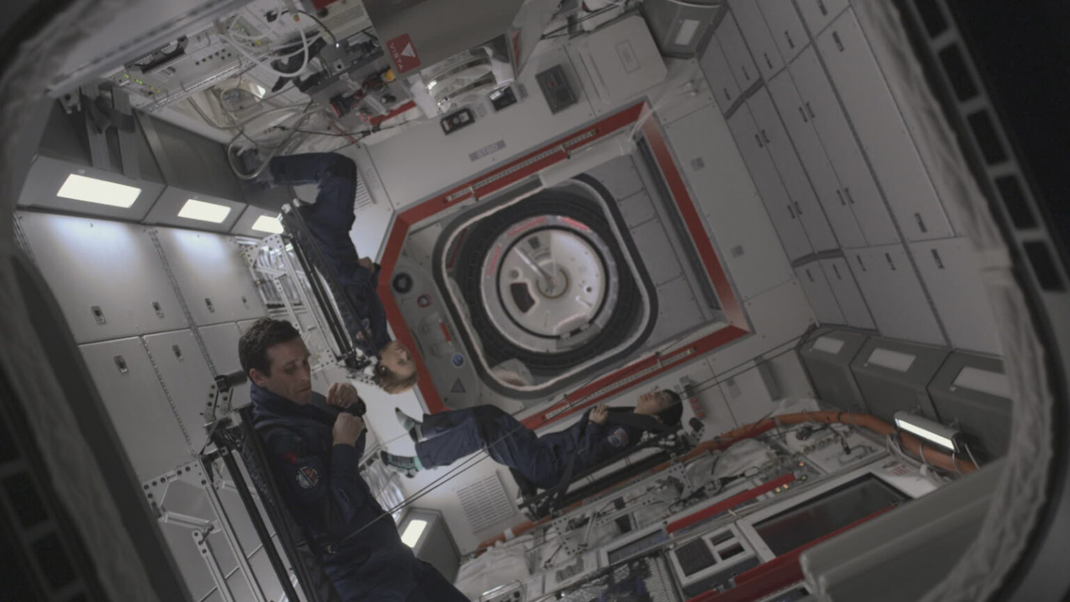 Three astronauts float inside a spacecraft module, with one holding onto a handrail. The interior is filled with equipment, controls, and a central hatch.