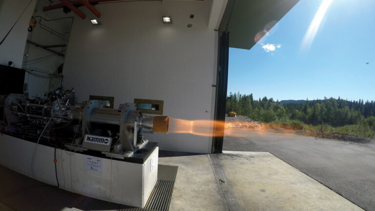 A rocket engine is undergoing a test firing, releasing flames and exhaust gases in an outdoor facility surrounded by a forest and clear sky.