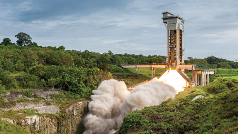 A rocket is launching from a platform, emitting bright flames and smoke, surrounded by green vegetation under a cloudy sky.