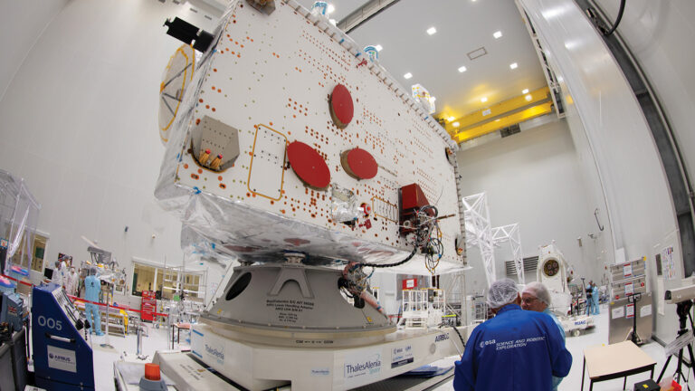 Technicians work on a large satellite in a cleanroom environment, surrounded by equipment and tools, under bright lights and a high ceiling.