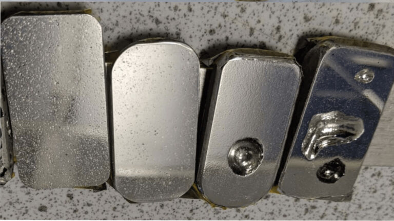 Five metallic blocks with different shapes and surface textures are arranged side by side on a speckled surface.