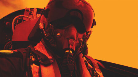 Close-up of a pilot wearing a helmet and oxygen mask in a cockpit with an orange-tinted background.