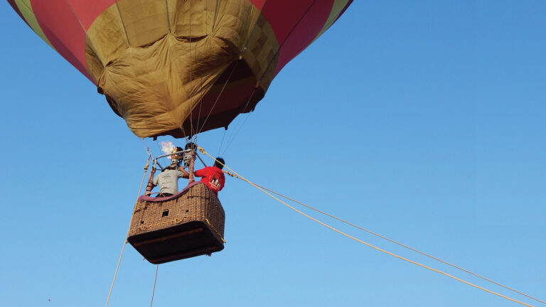 A person in a red jacket stands in the wicker basket of a colorful hot air balloon against a clear blue sky.