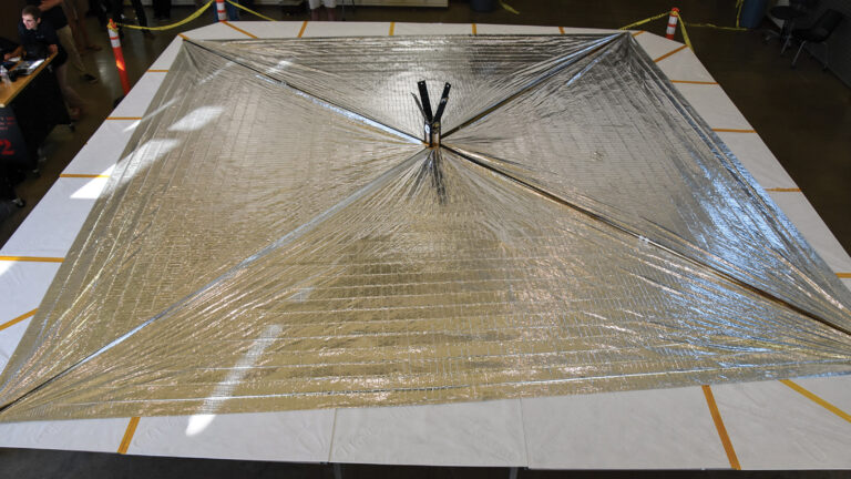 A large, reflective, square-shaped solar sail is displayed indoors on a flat surface. The sail is supported by a central pole and is bordered by white material with yellow tape.