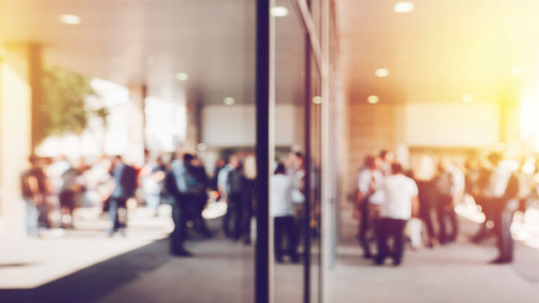 A crowd of people standing and walking in an outdoor area near a building, with a glass wall reflecting part of the scene, and sunlight creating a warm tone.