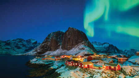 Small red cabins sit along a snowy shoreline beneath the Northern Lights, with a large rocky mountain in the background and a starry night sky above.