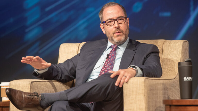 A man in a suit and tie sits on a beige armchair, gesturing with his right hand while talking. A black coffee tumbler is on a table next to him, and a blue, abstract background is behind him.